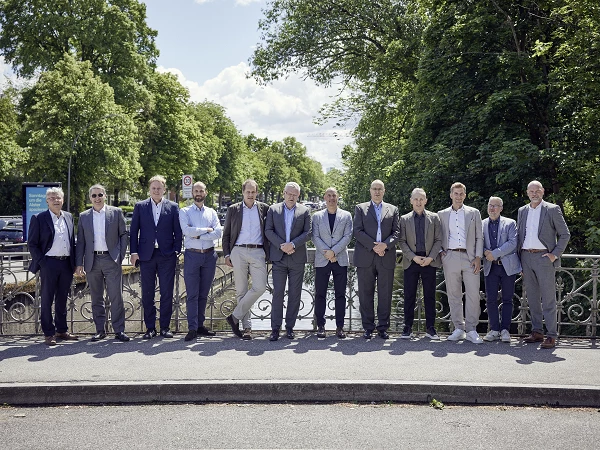 Eine Gruppe von zwölf Männern in formeller Kleidung steht auf einer Brücke über einem Fluss, umgeben von Bäumen und grünem Laub. Der Himmel ist blau mit einigen Wolken.