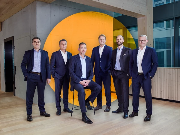Sechs Männer in formeller Kleidung stehen und sitzen in einem modernen Büro mit großen Fenstern und sichtbarem orangefarbenem Element im Hintergrund.