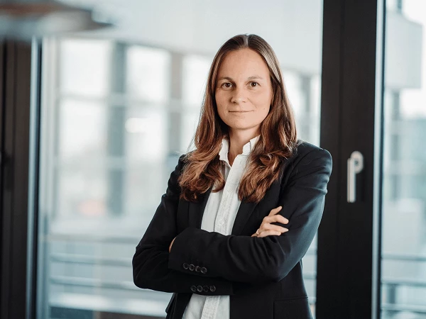 Eine Frau mit langen, braunen Haaren steht mit verschränkten Armen vor einem Fenster in einem modernen Büro. Sie trägt ein schwarzes Sakko und ein weißes Hemd und schaut direkt in die Kamera.