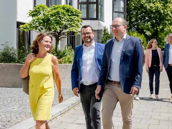 Eine Gruppe von fünf Personen läuft entspannt auf einem Gehweg entlang einer lichtdurchfluteten Straße, umgeben von modernen Gebäuden und Bäumen. Drei Personen im Vordergrund, eine Frau in einem gelben Kleid und zwei Männer in Anzügen, lachen und unterhalten sich, während zwei weitere Personen im Hintergrund an der Seite stehen.