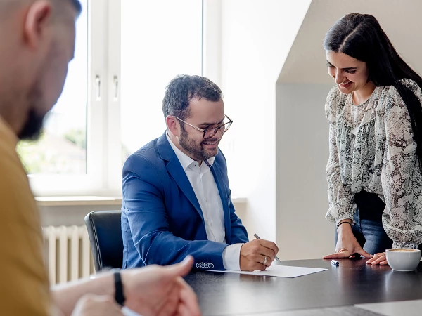 Ein Mann in einem blauen Anzug sitzt an einem Tisch und schreibt auf einem Dokument. Eine Frau steht neben ihm und schaut aufmerksam auf das Papier, während eine weitere Person im Hintergrund die Szene beobachtet. In einer Schale auf dem Tisch sind Snacks zu sehen.