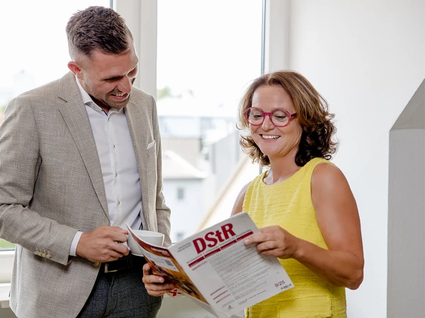 Ein Mann in einem hellen Anzug und eine Frau in einem gelben Kleid stehen an einem Fenster und lächeln, während sie gemeinsam eine Zeitschrift mit dem Titel "DSR" betrachten.