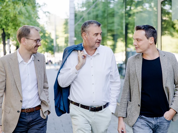 Drei Männer in eleganter Kleidung gehen lächelnd nebeneinander auf einem Gehweg, umgeben von Bäumen und Glasgebäuden. Der Mann in der Mitte trägt einen blauen Blazer über der Schulter.
