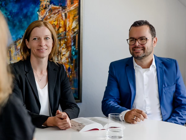 Zwei Personen in formeller Kleidung sitzen an einem Tisch, lächeln und unterhalten sich, während eine dritte Person, die nicht im Bild zu sehen ist, ihnen gegenüber sitzt. Im Hintergrund ist ein farbenfrohes Gemälde zu erkennen. Auf dem Tisch steht ein Glas Wasser und ein aufgeschlagenes Buch.