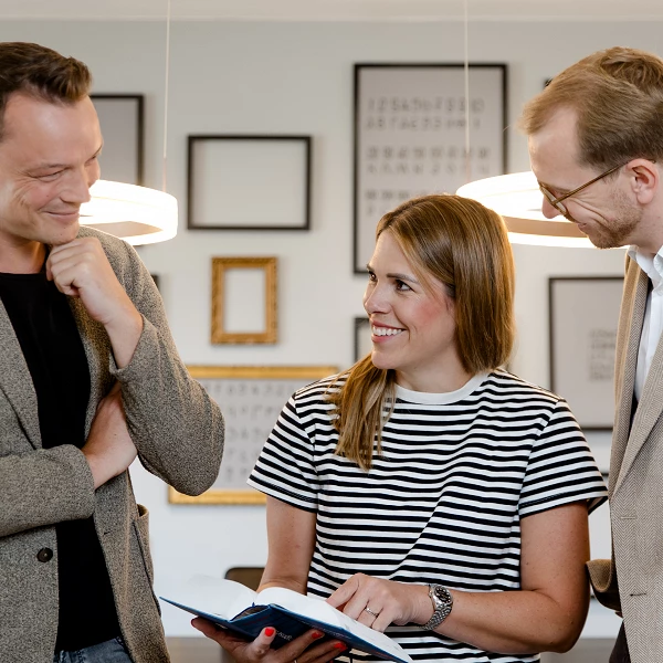 Drei Personen in einem modernen Raum stehen zusammen und betrachten ein Buch, während sie miteinander lächeln und sich unterhalten. Die Frau in der Mitte trägt ein gestreiftes T-Shirt, während die beiden Männer daneben Blazer anhaben. Im Hintergrund sind Bilderrahmen an der Wand zu sehen.