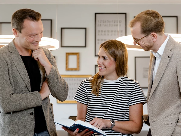 Drei Personen in einem modernen Raum stehen zusammen und betrachten ein Buch, während sie miteinander lächeln und sich unterhalten. Die Frau in der Mitte trägt ein gestreiftes T-Shirt, während die beiden Männer daneben Blazer anhaben. Im Hintergrund sind Bilderrahmen an der Wand zu sehen.
