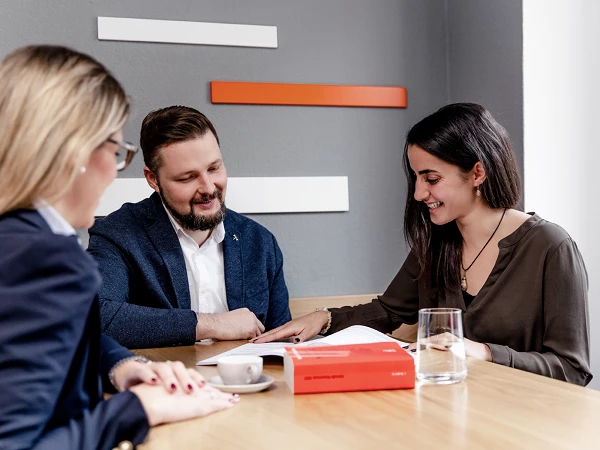 Drei Personen sitzen an einem Tisch in einem modernen Raum, zwei Frauen und ein Mann, die an einem Dokument arbeiten. Eine Frau zeigt auf das Dokument, während der Mann aufmerksam zuhört. Auf dem Tisch steht ein Kaffeebecher und ein klares Glas Wasser. Im Hintergrund sind farbige Akzentwände zu sehen.