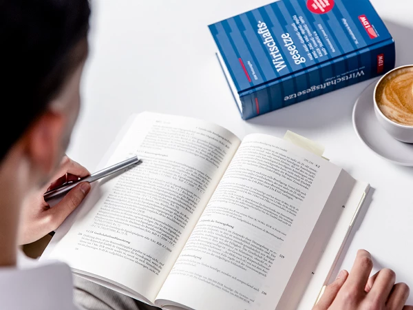 Eine Person sitzt an einem Tisch und liest ein Buch mit dem Titel „Wirtschaftsgesetze“, während sie mit einem Stift Notizen macht. Neben dem Buch steht eine Tasse mit Getränk.