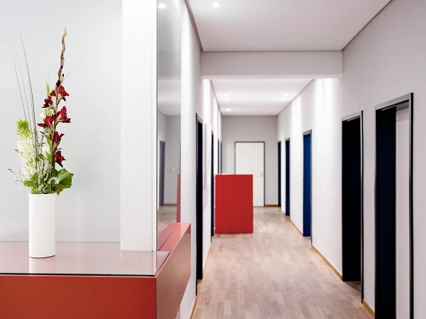 Ein moderner Flur mit hellen Wänden und Holzfußboden, an dessen Ende eine geschlossene Tür zu sehen ist. Auf der linken Seite stehen mehrere schwarze Türen, während rechts ein blühendes Arrangement in einer weißen Vase auf einer rot lackierten Theke platziert ist.