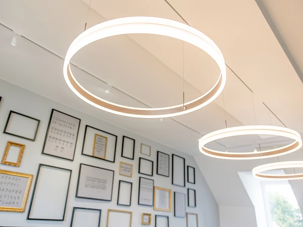 Hängende, runde Leuchten aus Lichtscheiben sind im oberen Bereich des Bildes zu sehen, während im Hintergrund eine Wand mit verschiedenen gerahmten Schriftstücken und Texten in unterschiedlichen Rahmen dekoriert ist.