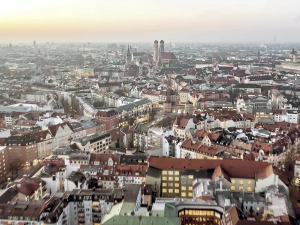 Blick auf die Stadt München bei Sonnenuntergang, mit einer Vielzahl von Dächern in unterschiedlichen Farben, historischen Gebäuden und einer weitläufigen urbanen Landschaft, die in der Ferne zu sehen ist.