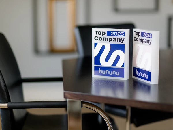 Zwei Auszeichnungen „Top Company 2024“ und „Top Company 2025“ von kununu stehen auf einem modernen Konferenztisch, während im Hintergrund Stühle und ein minimalistisches Raumdesign sichtbar sind.