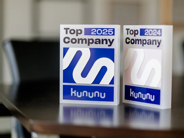 Zwei Auszeichnungen mit der Aufschrift "Top Company" für die Jahre 2024 und 2025 stehen auf einem Tisch. Die Auszeichnungen sind im gleichen Design und zeigen das Logo von kununu. Im Hintergrund sind verschwommen Möbel zu sehen.