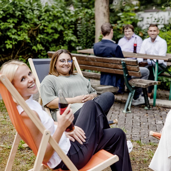 Eine Gruppe von Menschen sitzt entspannt in einem Garten, zwei Frauen in Liegestühlen im Vordergrund lächeln und halten Getränke, während im Hintergrund weitere Personen an einem Tisch miteinander sprechen.
