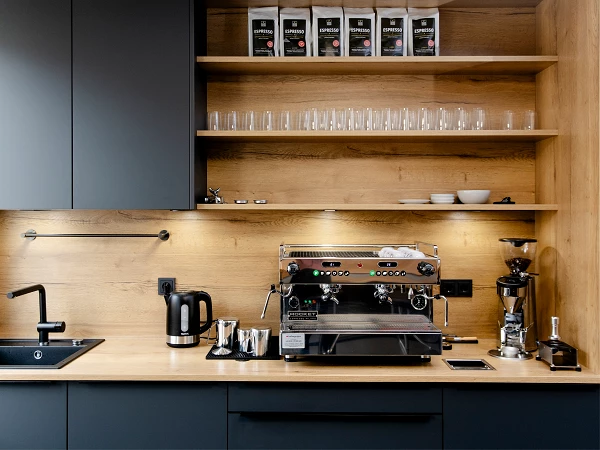 Eine moderne Kaffeeküche mit einer professionellen Espressomaschine, einer Kaffeemühle, einem Wasserkocher und Gläsern auf einem Holzregal, alles in eleganten, dunklen Farbtönen.