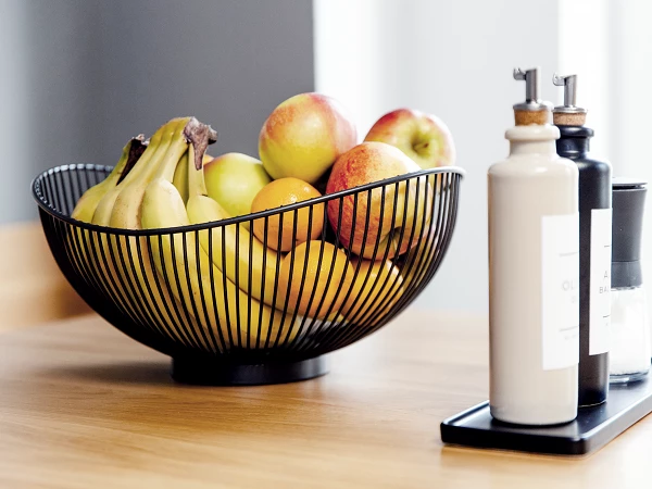 Ein schwarzer Drahtkorb mit Äpfeln und Bananen steht auf einem Holztisch, daneben befinden sich drei Flaschen mit verschiedenen Flüssigkeiten und einer Gewürzmühle auf einem schmalen Tablett.