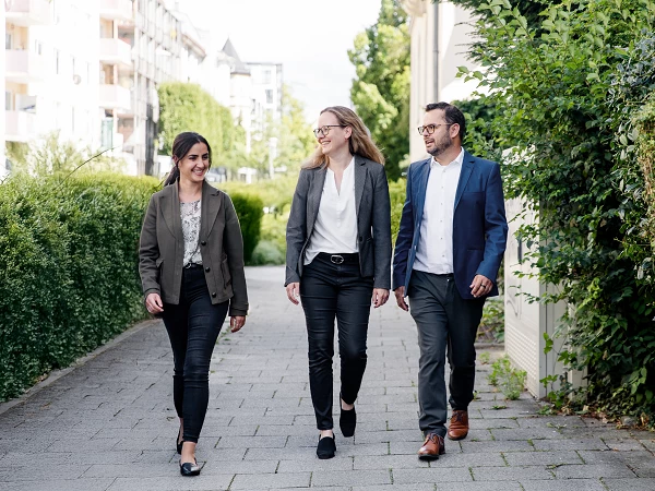 Drei Personen in formeller Kleidung gehen auf einem Gehweg entlang, umgeben von Grünanlagen und Gebäuden. Sie wirken freundlich und selbstbewusst.