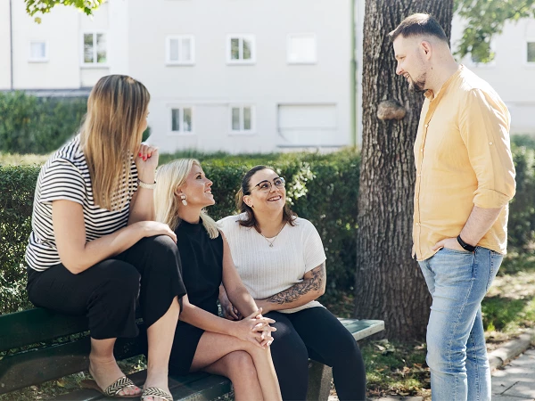 Vier Personen unterhalten sich entspannt auf einer Bank in einem Park. Zwei Frauen sitzen auf der Bank, eine dritte steht neben ihnen, während ein Mann frontal gegenüber steht. Alle wirken freundlich und amüsiert, umgeben von Bäumen und einer hellen, sonnigen Umgebung.