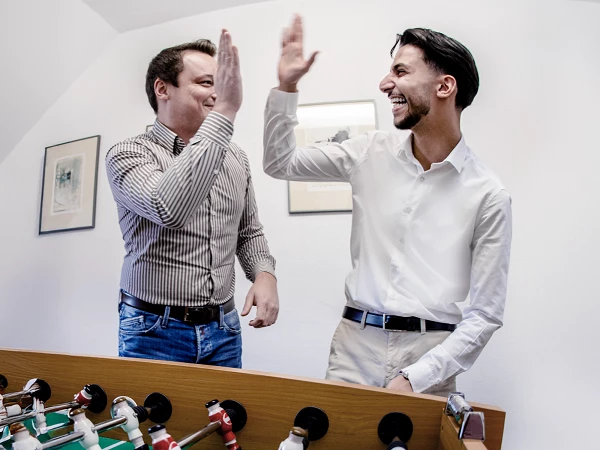 Zwei Männer stehen an einem Tischkicker und geben sich einen High-Five, während sie lächeln und offensichtlich Spaß haben.