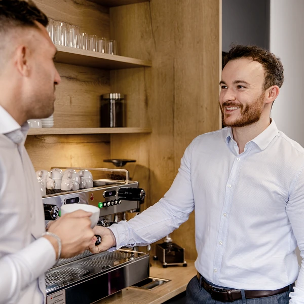 Zwei Männer in eleganter Kleidung stehen an einer Kaffeemaschine in einer modernen Küche, während einer von ihnen eine Tasse Kaffee in der Hand hält und lächelt.