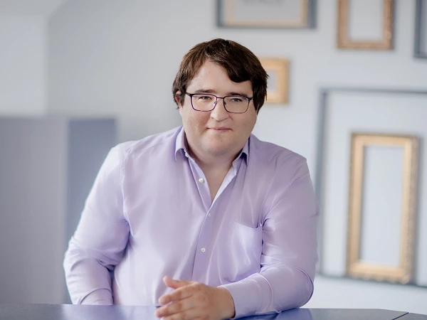 Ein Mann mit brünettem, lockigem Haar und Brille sitzt an einem Tisch, trägt ein lilanes Hemd und schaut direkt in die Kamera. Im Hintergrund sind einige eingerahmte Bilder an der Wand sichtbar.