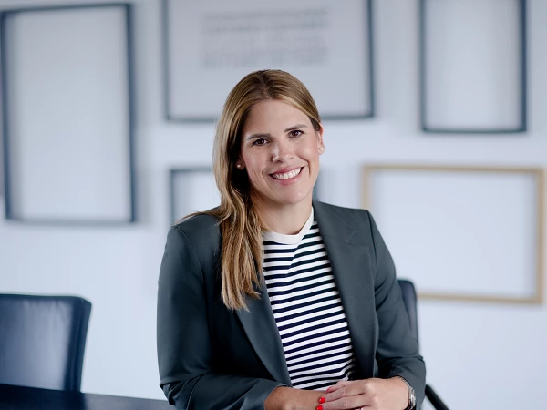 Eine lächelnde Frau mit langen, hellen Haaren sitzt an einem Schreibtisch, trägt ein gestreiftes Oberteil und einen grauen Blazer, während sie in einem modernen Büro mit leeren Bilderrahmen im Hintergrund posiert.