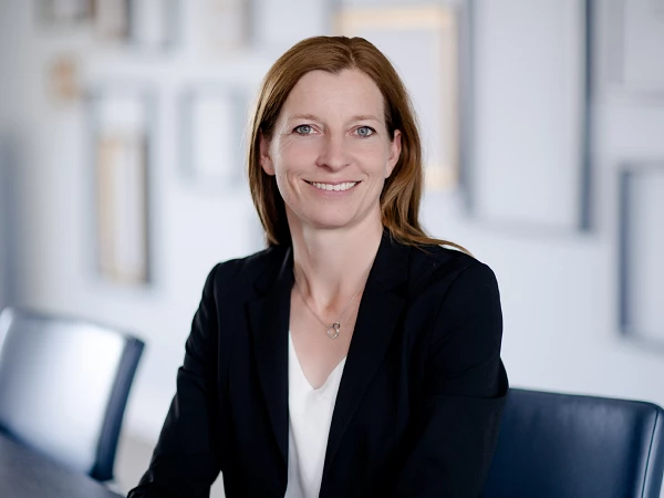 Eine Frau mit langen, braunen Haaren sitzt an einem Tisch in einem modernen Büroraum und lächelt freundlich in die Kamera. Sie trägt einen schwarzen Blazer und ein weißes Oberteil. Im Hintergrund sind verschwommene, eingerahmte Bilder an der Wand zu sehen.