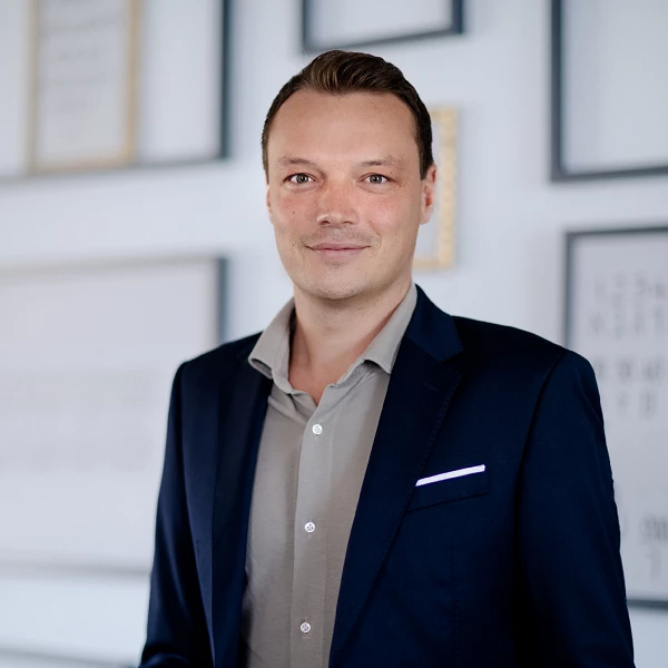 Ein Mann in einem dunklen Anzug mit hellen Hemd steht vor einem Wandarrangement aus Bilderrahmen und Grafiken.