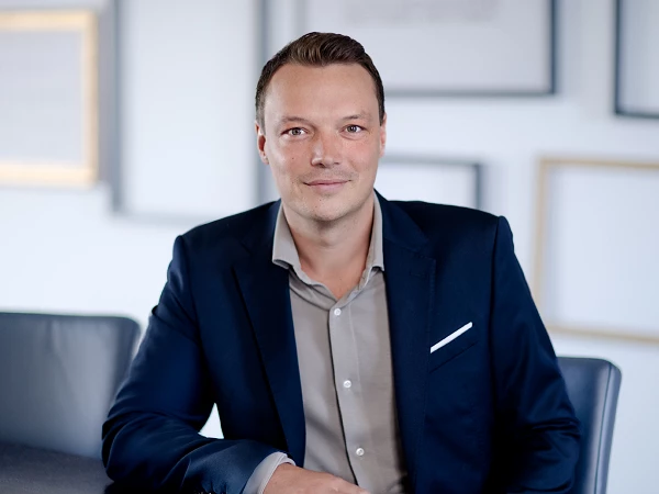 Ein Mann in einem dunklen Anzug sitzt am Tisch in einem modernen Raum mit gerahmten Bildern im Hintergrund und lächelt in die Kamera.