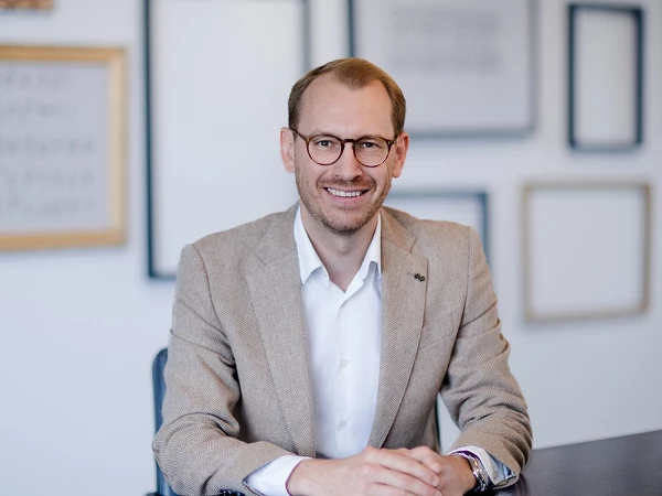 Ein Mann mit Brille sitzt lächelnd an einem Tisch, gekleidet in einem hellen Sakko und einem weißen Hemd. Im Hintergrund sind mehrere rahmenlose Bilder an der Wand zu sehen.