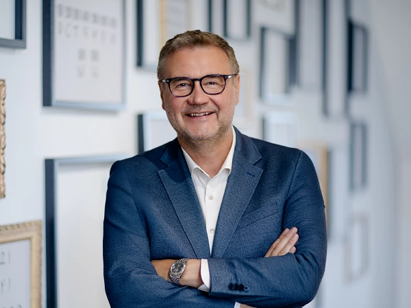 Ein Mann mit Brille, der in einem eleganten blauen Anzug und einem weißen Hemd gekleidet ist, steht lächelnd mit übergeschlagenen Armen vor einer Wand mit gerahmten Bildern und Texten.
