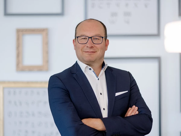 Ein Mann in einem blauen Anzug mit Brille steht selbstbewusst mit verschränkten Armen vor einer Wand mit gerahmten Bildern.