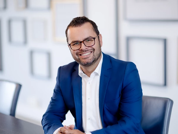 Ein lächelnder Mann mit Brille trägt einen blauen Anzug und sitzt an einem Tisch in einem modernen Büro mit Rahmen an der Wand im Hintergrund.