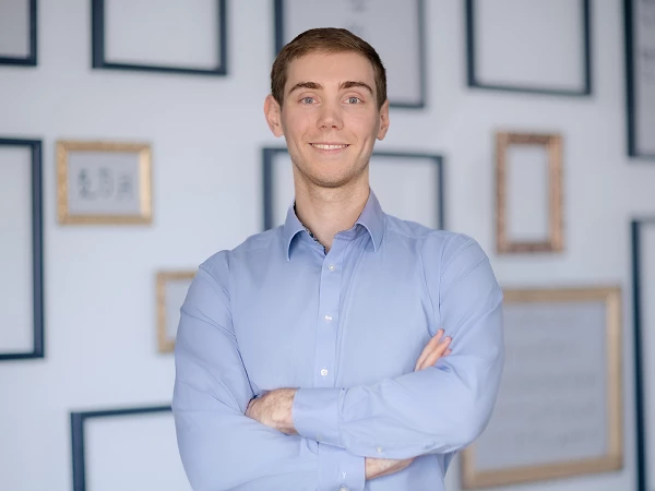 Ein junger Mann mit einem freundlichen Lächeln steht mit verschränkten Armen vor einer Wand mit mehreren Bilderrahmen. Er trägt ein hellblaues Hemd und präsentiert eine positive Ausstrahlung.