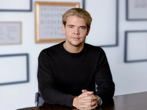 Ein junger Mann mit blonden Haaren trägt einen schwarzen Pullover und sitzt an einem Tisch, die Hände gefaltet. Im Hintergrund sind Bilderrahmen an der Wand sichtbar, die jedoch keine Bilder enthalten.