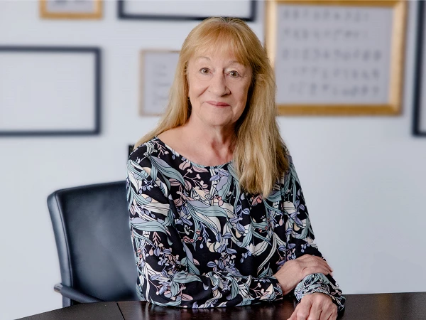 Eine lächelnde Frau mit langen blonden Haaren sitzt an einem Tisch, bekleidet mit einem blumenmusternden Oberteil. Im Hintergrund sind gerahmte Bilder an der Wand zu sehen.