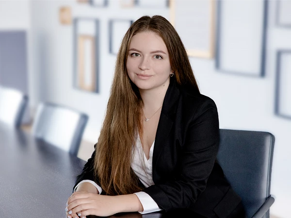 Eine junge Frau mit langen, glatten Haaren sitzt am Tisch in einem modern gestalteten Büro, trägt einen schwarzen Blazer und ein weißes Hemd und lächelt in die Kamera. Im Hintergrund sind verschwommene Bilderrahmen an der Wand zu sehen.