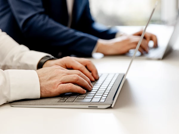 Zwei Personen sitzen an einem Tisch und tippen auf Laptops; eine Person in einem weißen Hemd und eine in einem dunkelblauen Anzug.