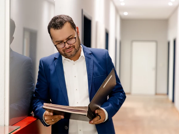 Ein Mann in einem blauen Anzug mit einer weißen Bluse steht in einem modernen Flur und schaut konzentriert in ein schwarzes Portfolio.