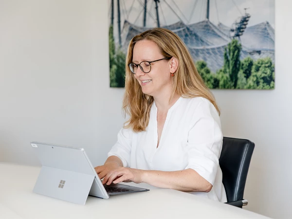 Eine Frau mit langen, blonden Haaren und Brille sitzt an einem Schreibtisch und arbeitet konzentriert an einem Laptop. Im Hintergrund ist ein unscharfes Sportstadion sichtbar.
