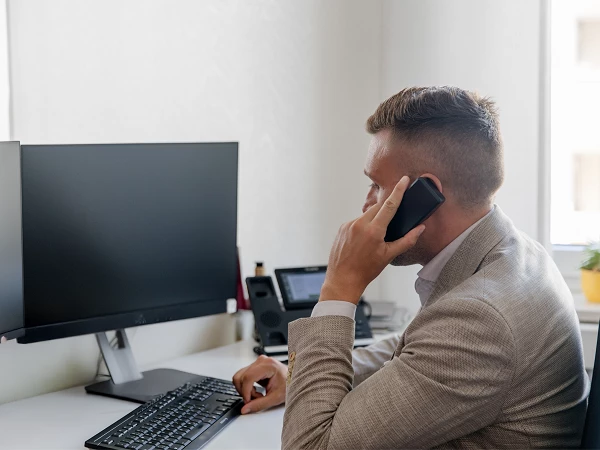 Ein Mann in einem hellen Anzug sitzt an einem Schreibtisch, hält ein Smartphone am Ohr und spricht, während er auf einen Computerbildschirm und seine Tastatur schaut. Auf dem Schreibtisch sind weitere Bürogeräte und eine kleine Pflanze zu sehen.