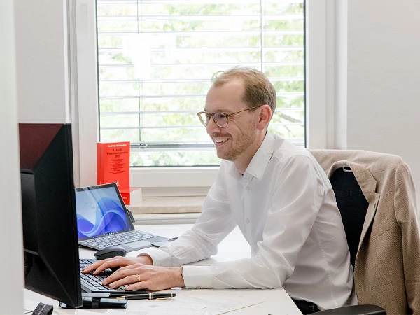 Ein Mann mit Brille sitzt an einem Schreibtisch und arbeitet an zwei Computern, während er lächelt. Hinter ihm ist ein Fenster mit Jalousien und ein rotes Informationsblatt zu sehen.