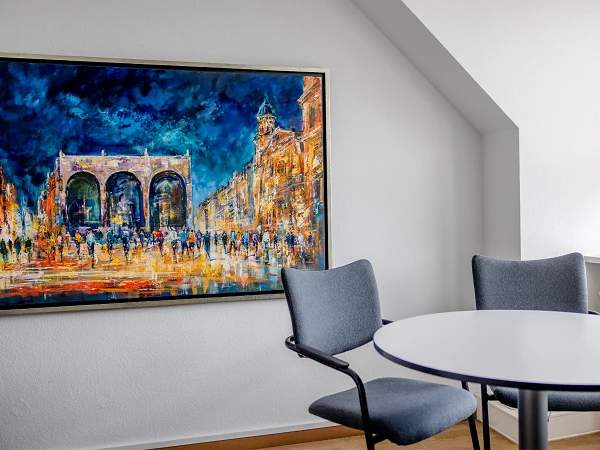 Ein farbenfrohes Gemälde eines Stadtbildes mit einem historischen Tor, das von Menschen in einer belebten Straße umgeben ist, hängt an einer Wand in einem modernen Raum mit einem runden Tisch und zwei Stühlen.