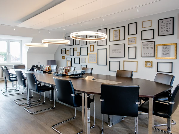 Ein moderner Besprechungsraum mit einem großen, rechteckigen Tisch und schwarzen Stühlen, umgeben von einer Wand mit verschiedenen gerahmten Bildern und Texten in unterschiedlichsten Rahmen. An der Decke hängen stilvolle, runde Leuchten.