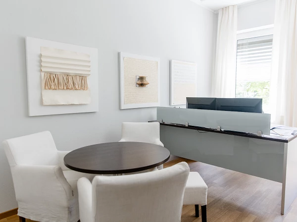 Ein moderner Büroraum mit einem grauen Schreibtisch, zwei weißen Sesseln und einem runden Tisch. An der Wand hängen drei Kunstwerke in neutralen Farben, und große Fenster lassen viel Tageslicht herein.