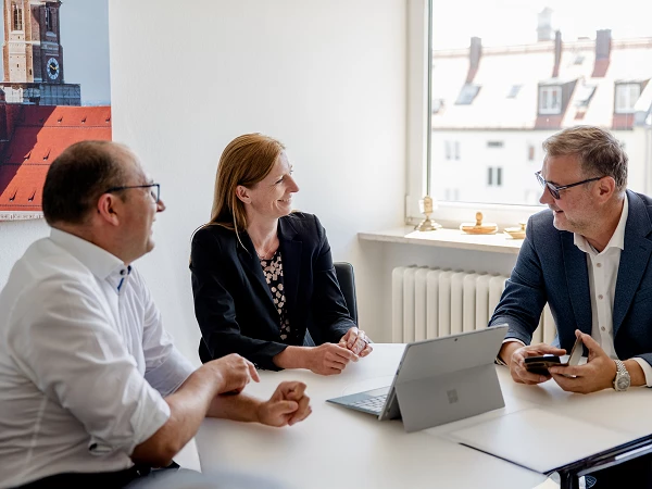 Drei Personen sitzen an einem Tisch in einem modernen Büro, während sie an einem Meeting teilnehmen; eine Frau in schwarzer Jacke lächelt, während die beiden Männer, einer mit einer Brille und der andere mit einem Tablet, aktiv mit ihr sprechen. Im Hintergrund sind Fenster mit Tageslicht und eine Stadtansicht sichtbar.