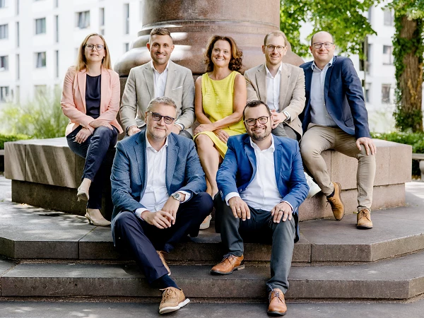 Eine Gruppe von sieben Personen sitzt und steht auf einer Steinbank in einem begrünten Außenbereich. Die Frauen tragen helle Blusen und Kleider, während die Männer Anzüge oder Blazer in unterschiedlichen Farben tragen. Im Hintergrund sind moderne Gebäude und Bäume zu sehen.