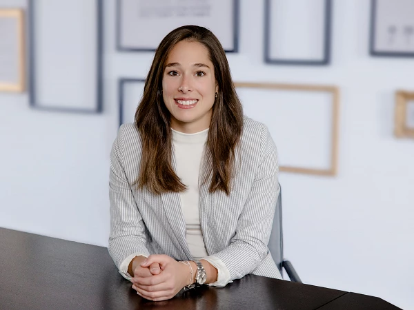 Eine lächelnde Frau mit langen, braunen Haaren sitzt an einem Tisch und trägt einen Streifenanzug, umgeben von einem minimalistischen Hintergrund mit Bilderrahmen.