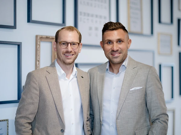 Zwei Männer stehen lächelnd nebeneinander vor einer Wand mit verschiedenen gerahmten Bildern und Grafiken. Beide tragen elegante Anzüge und haben eine freundliche Ausstrahlung.