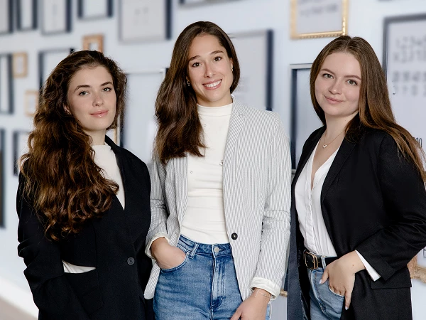 Drei junge Frauen stehen lächelnd in einem modern gestalteten Raum mit gerahmten Bildern im Hintergrund. Die erste Frau hat lange, lockige Haare und trägt einen schwarzen Blazer. Die zweite Frau hat mittellange, glatte Haare und trägt einen grauen Blazer. Die dritte Frau hat braune Haare und trägt ebenfalls einen schwarzen Blazer.